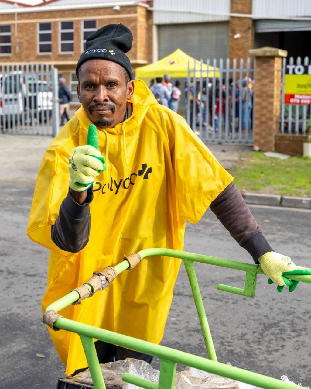 A WasteGo Green collector in a yellow Polyco poncho giving a thumbs up while pushing the green recycling collections trolley