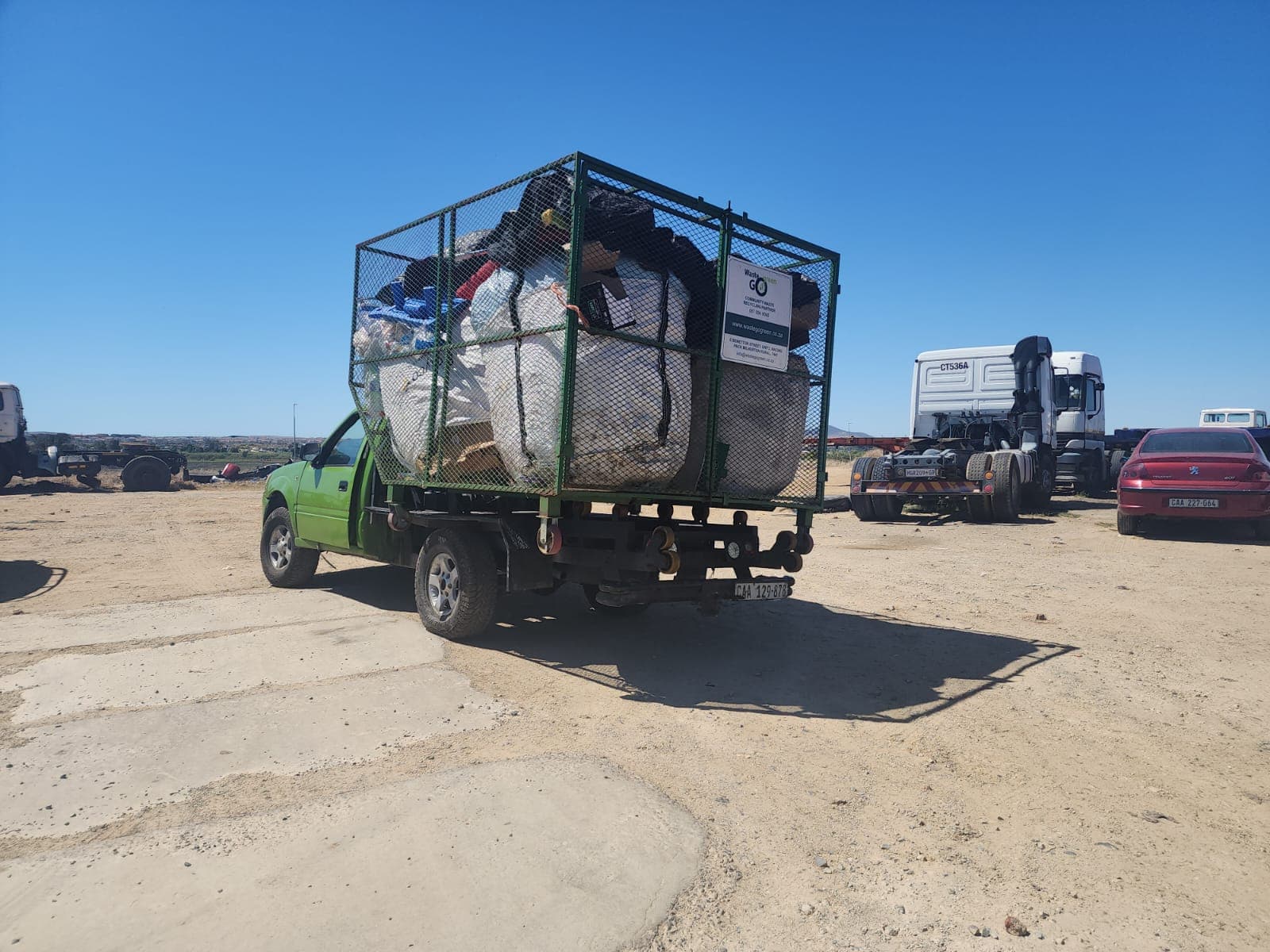 A green bakkie with a cage of bulk bags on an open field with truck-tractor units in a Cape Town township