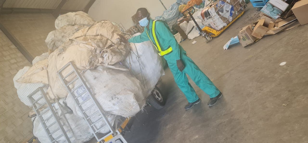 A worker in green overalls and a hi-vis vest moving bulk bags of recyclables with a pallet jack inside the warehouse
